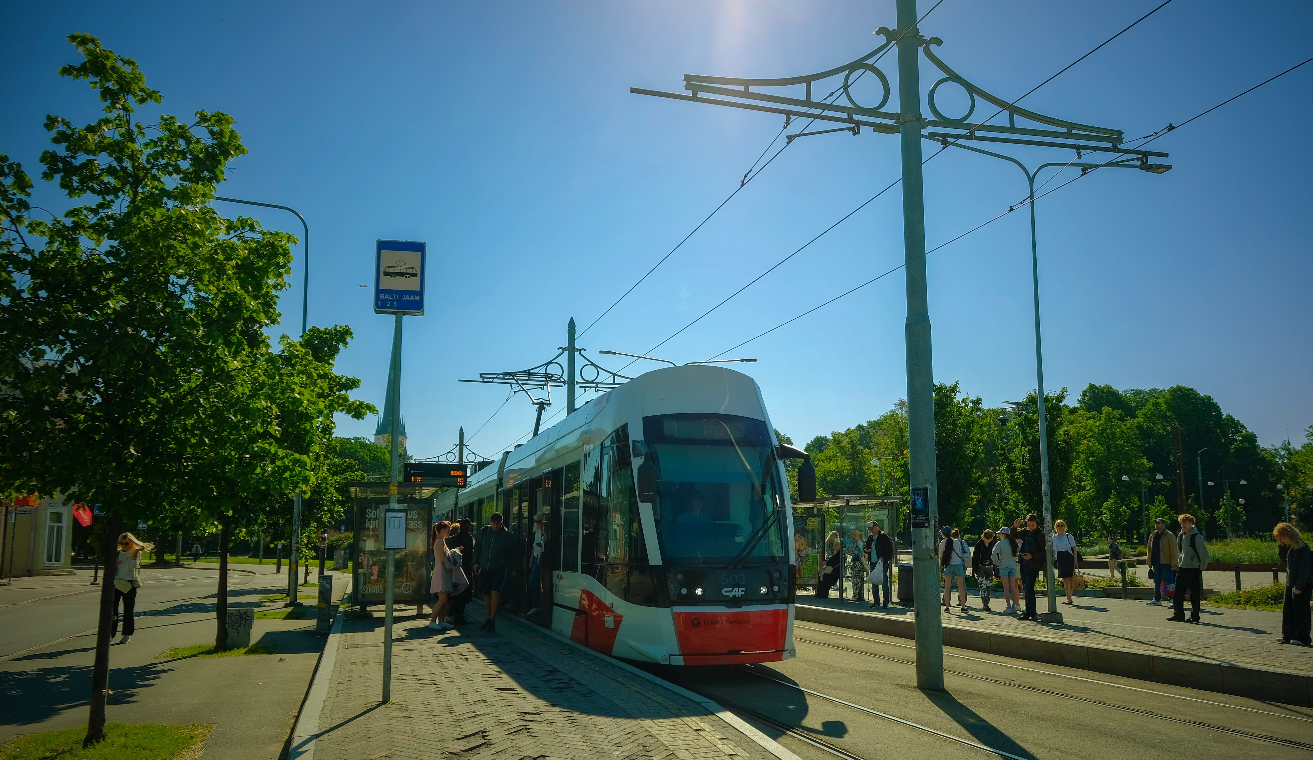 Kaasaegne tramm Balti Jaama trammipeatuses Tallinnas, reisijad ootavad p&auml;ikesepaistelisel p&auml;eval.