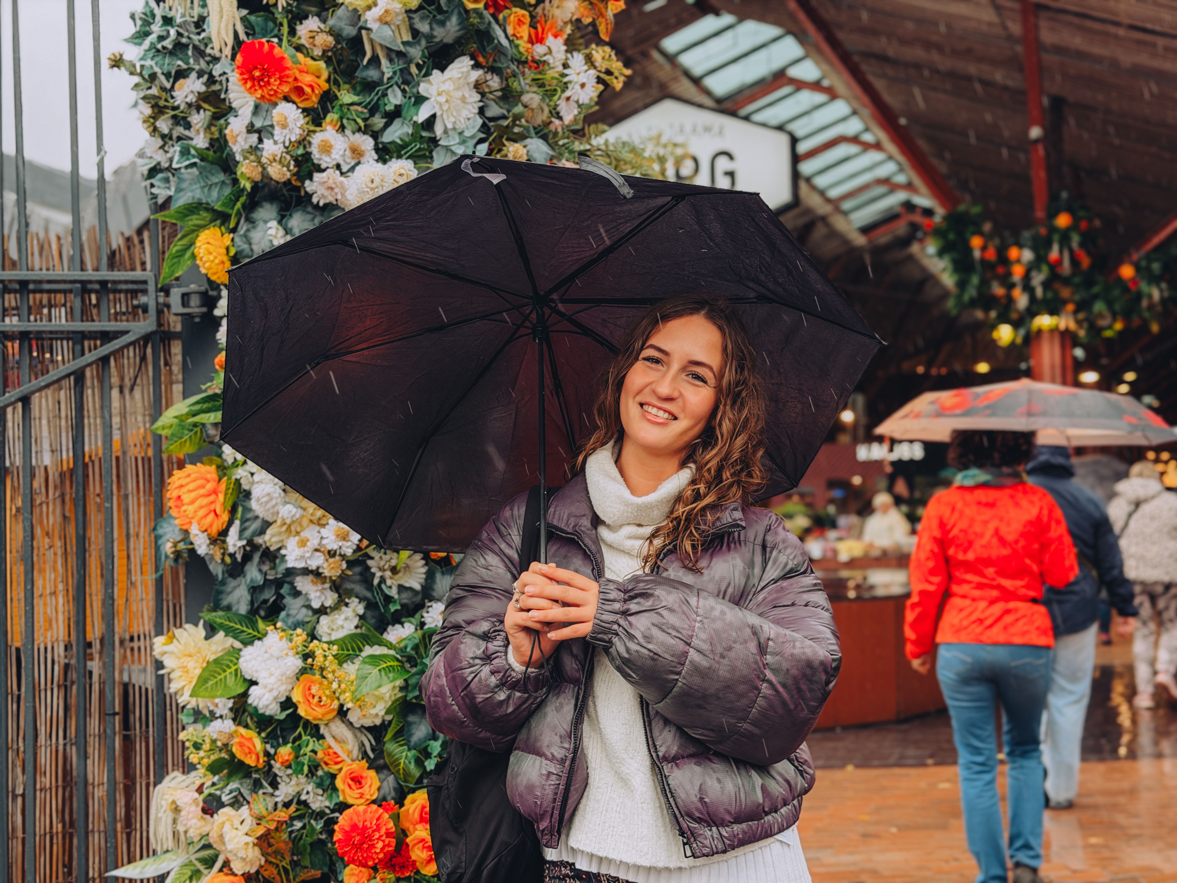 Leila smiles under a black umbrella at Balti Jaam Market in Tallinn, beside a colourful flower wall with market stalls behind her.