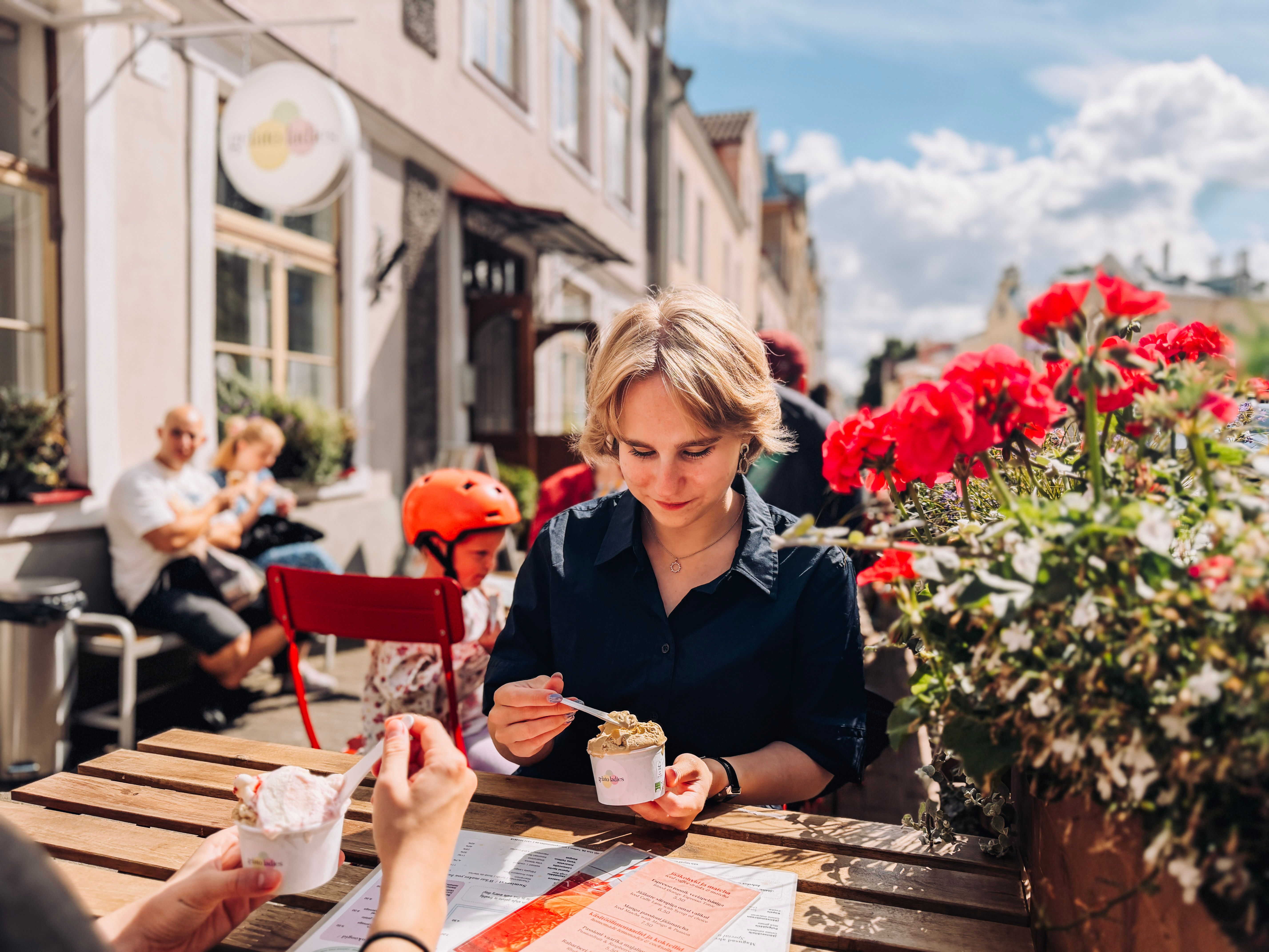 Carmen eats ice cream at a sunny caf&eacute; terrace in Tallinn Old Town beside red geraniums, with other guests in the background.