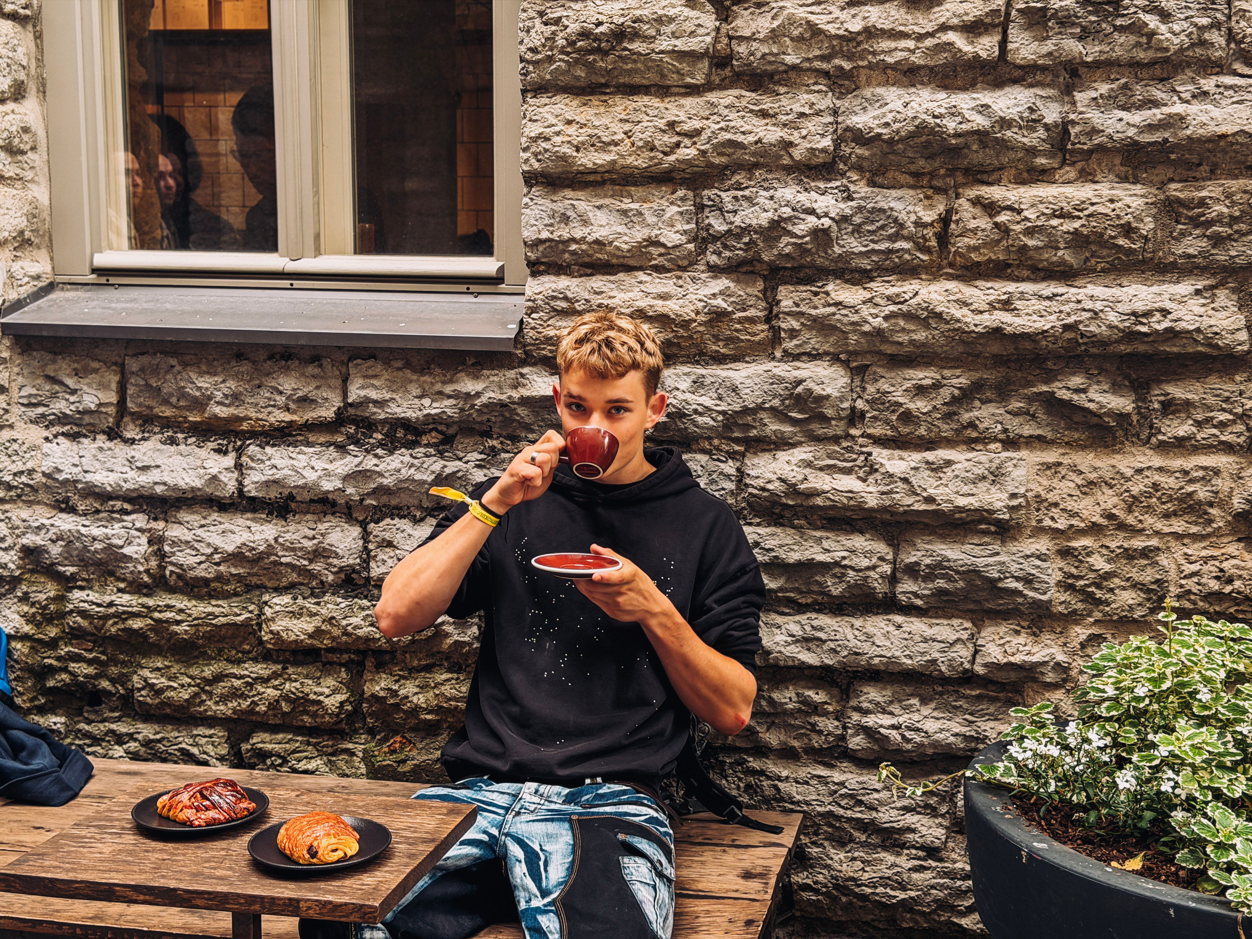 Frederik drinks coffee from a red cup in Tallinn&rsquo;s Rotermann Quarter, seated by a stone wall with two pastries on a wooden table.