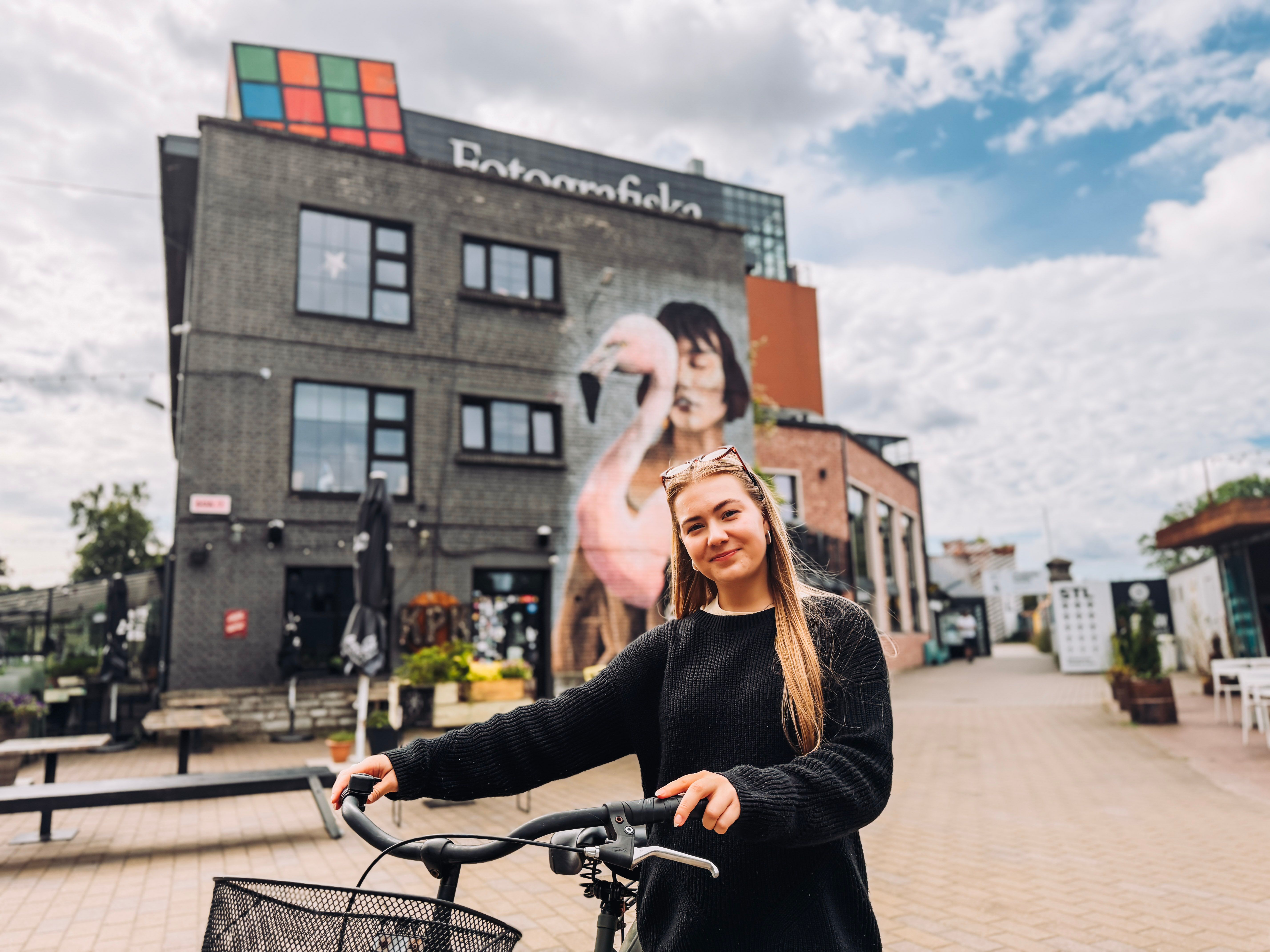 Liisa holds a bicycle outside Fotografiska Tallinn in Telliskivi, with a large flamingo mural covering the building behind her.