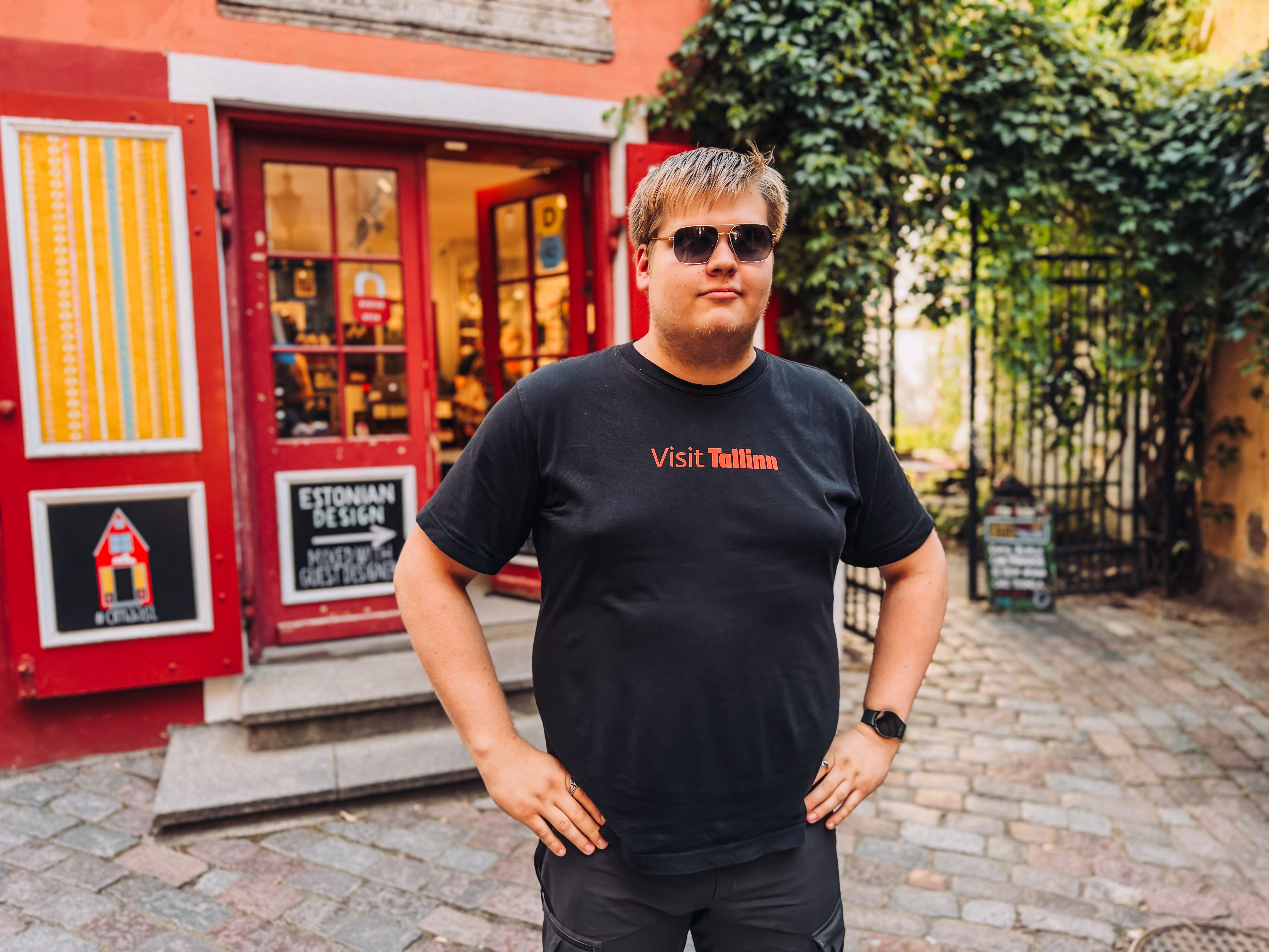 Sander stands on a cobblestone street in Tallinn Old Town wearing sunglasses and a black Visit Tallinn T-shirt by a red shop.