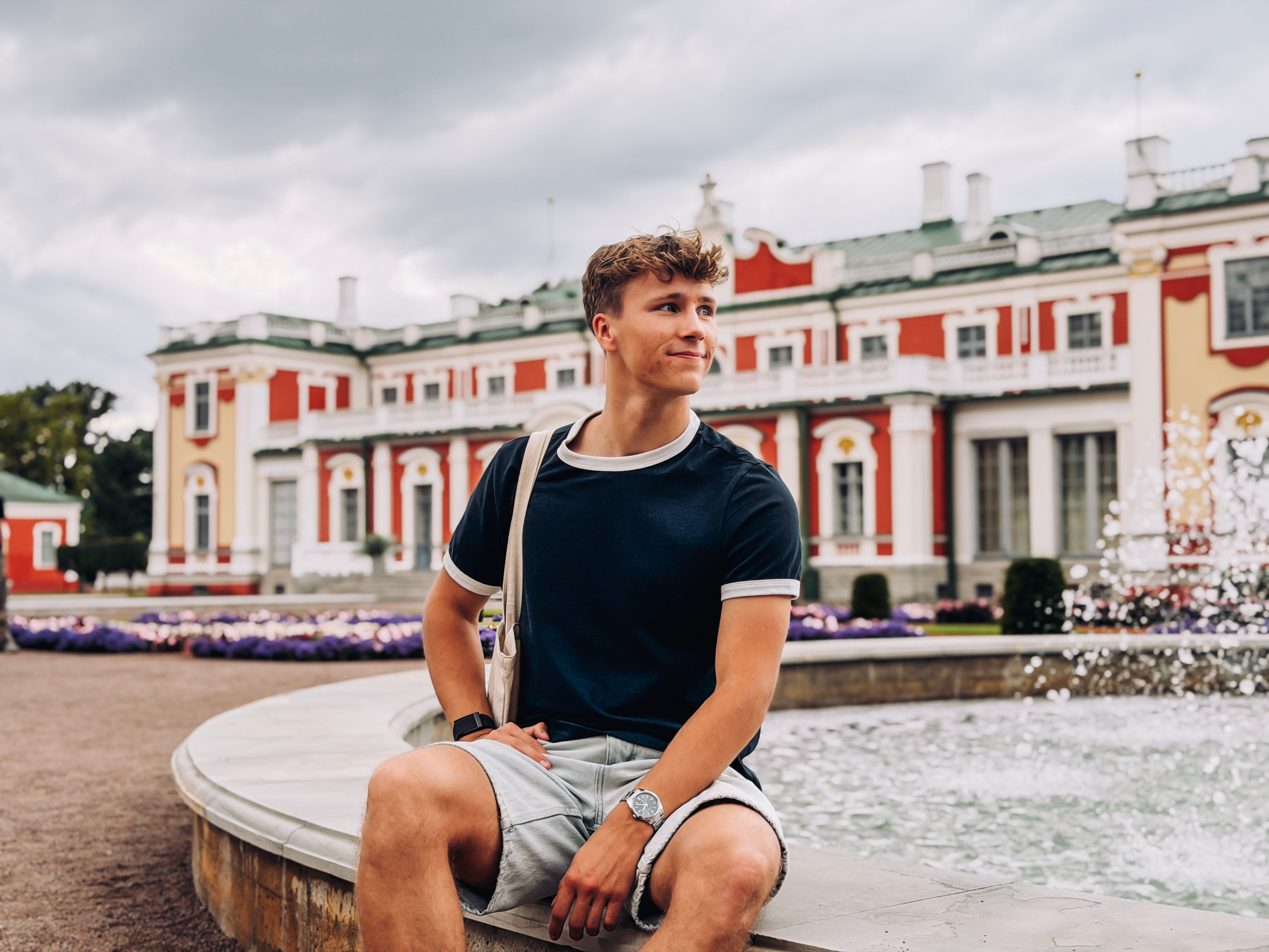 Karl-Kristjan sits beside a fountain in Kadriorg Park, with the red-and-white baroque Kadriorg Palace in Tallinn behind him.