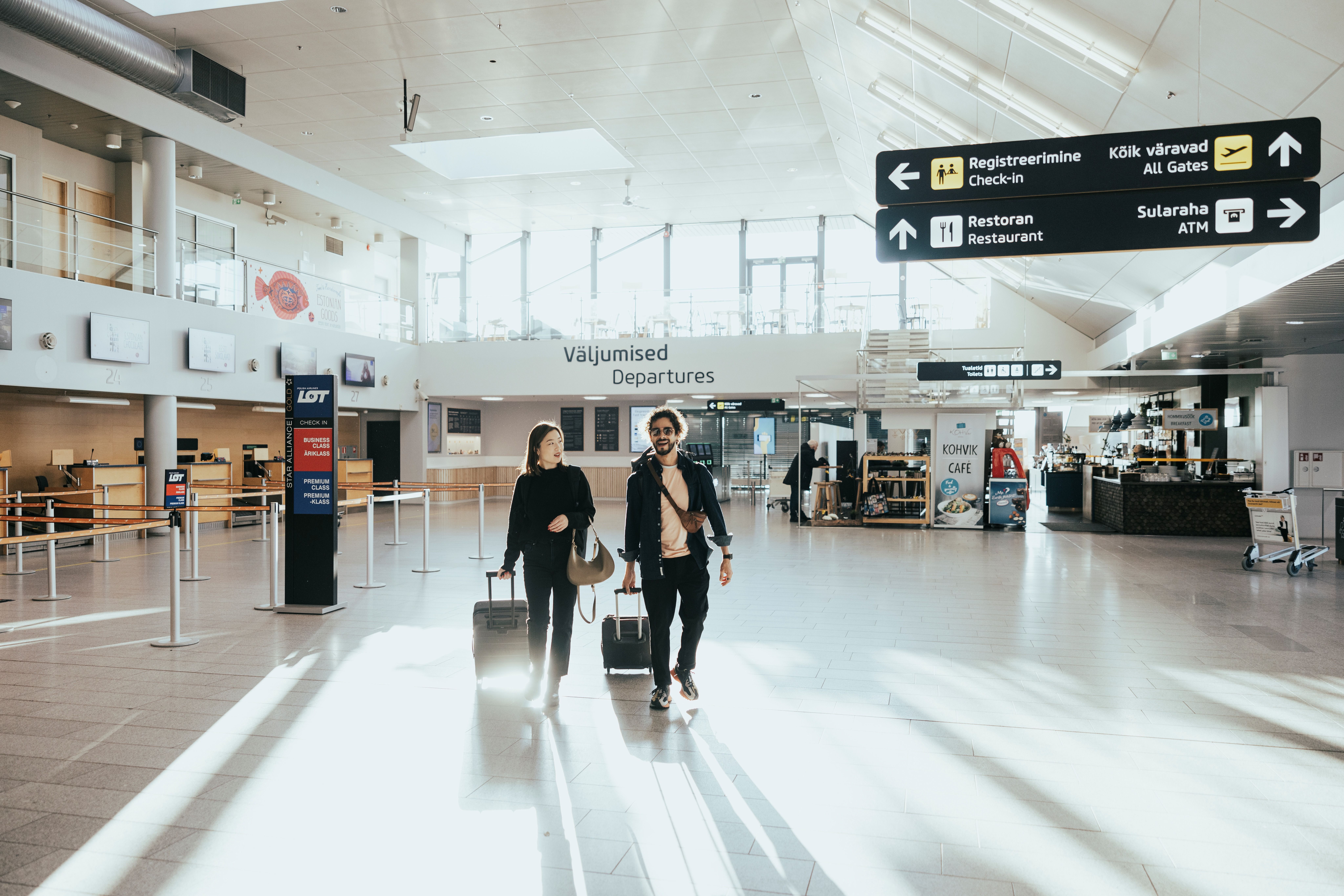 Two travellers with suitcases walk through Tallinn Airport departures hall, with check-in desks, a caf&eacute; and directional signs behind them.