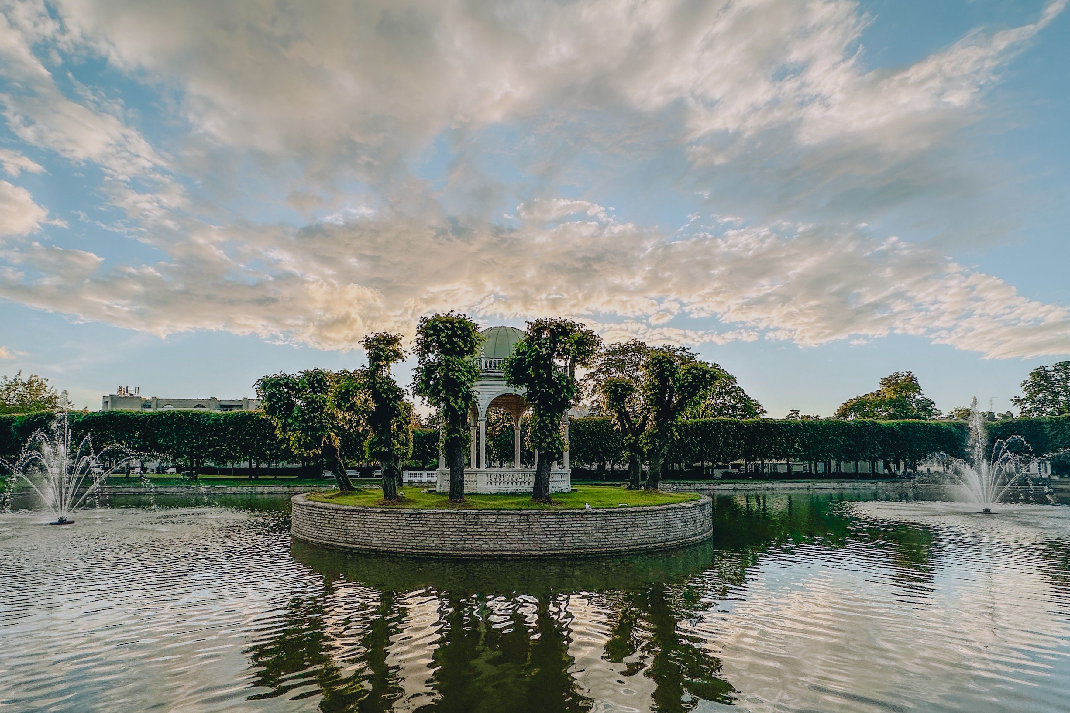 A scenic view of a pavilion on an island surrounded by fountains in the pond at Kadriorg Palace Park, with lush trees and a dramatic sky at sunset.