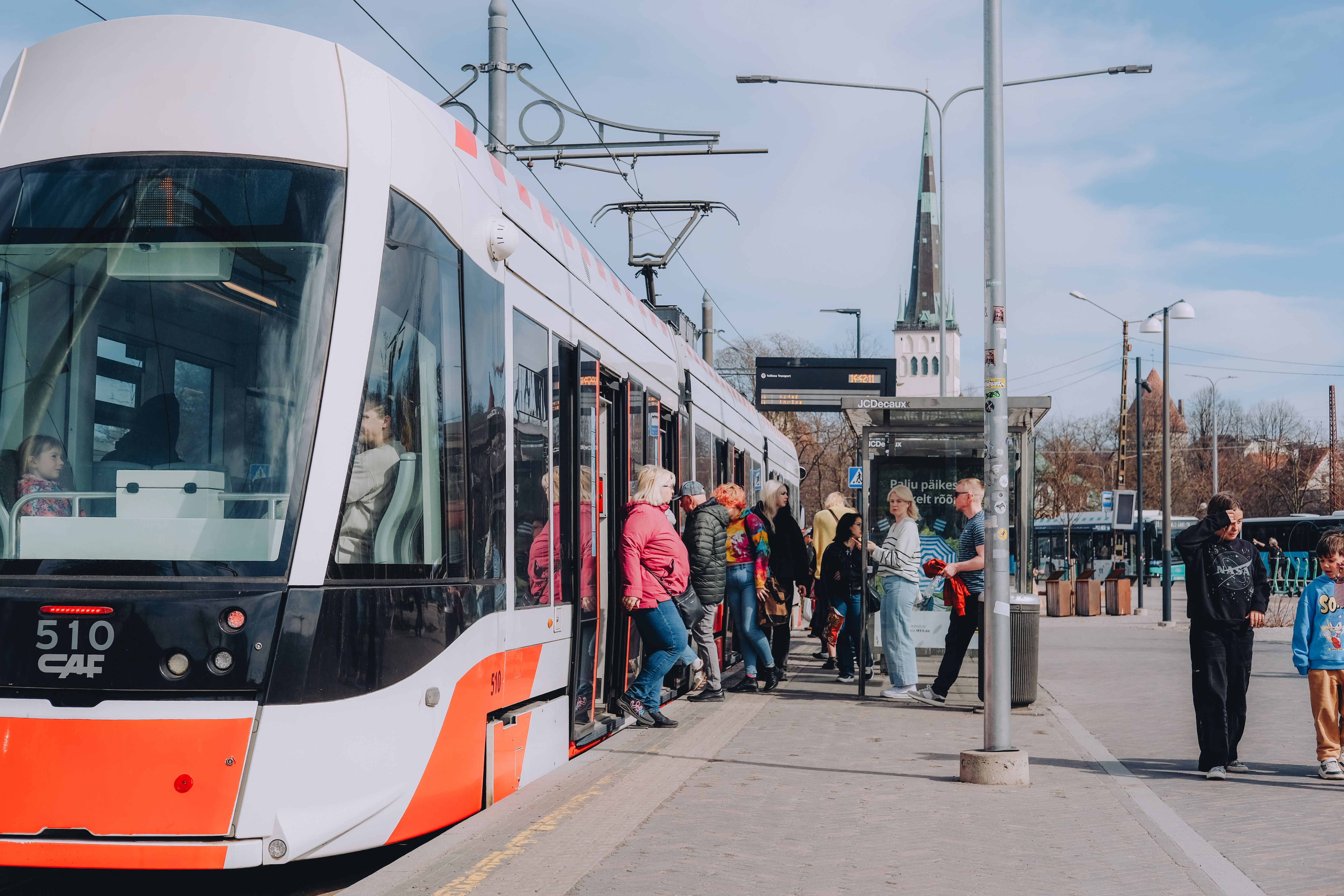 People boarding and exiting a modern red-and-white tram at a busy stop in central Tallinn, with St. Olaf’s Church visible in the background.