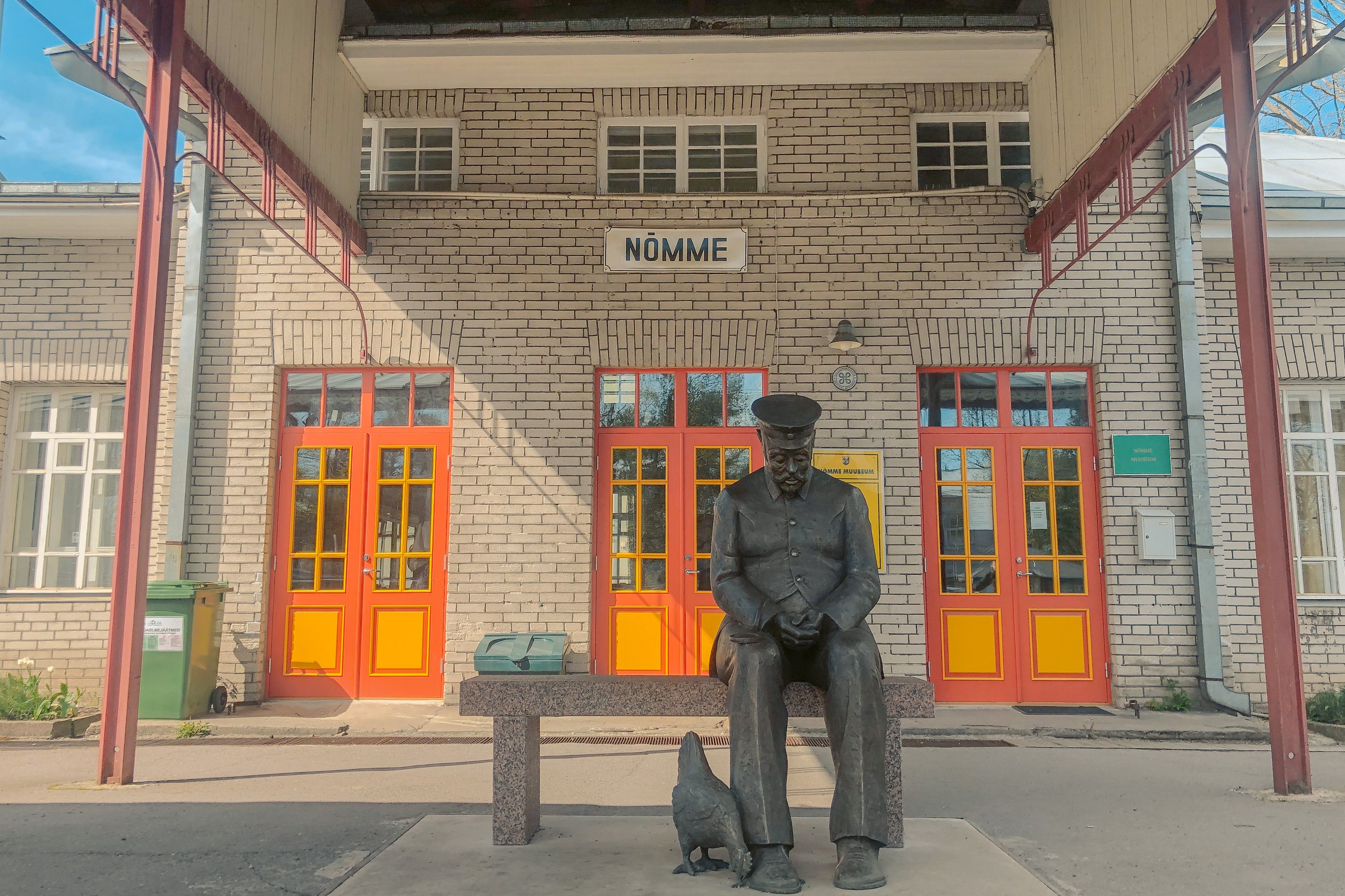 A view of Nõmme train station with its distinctive red and yellow doors and a statue of a station master sitting with a chicken at his feet.