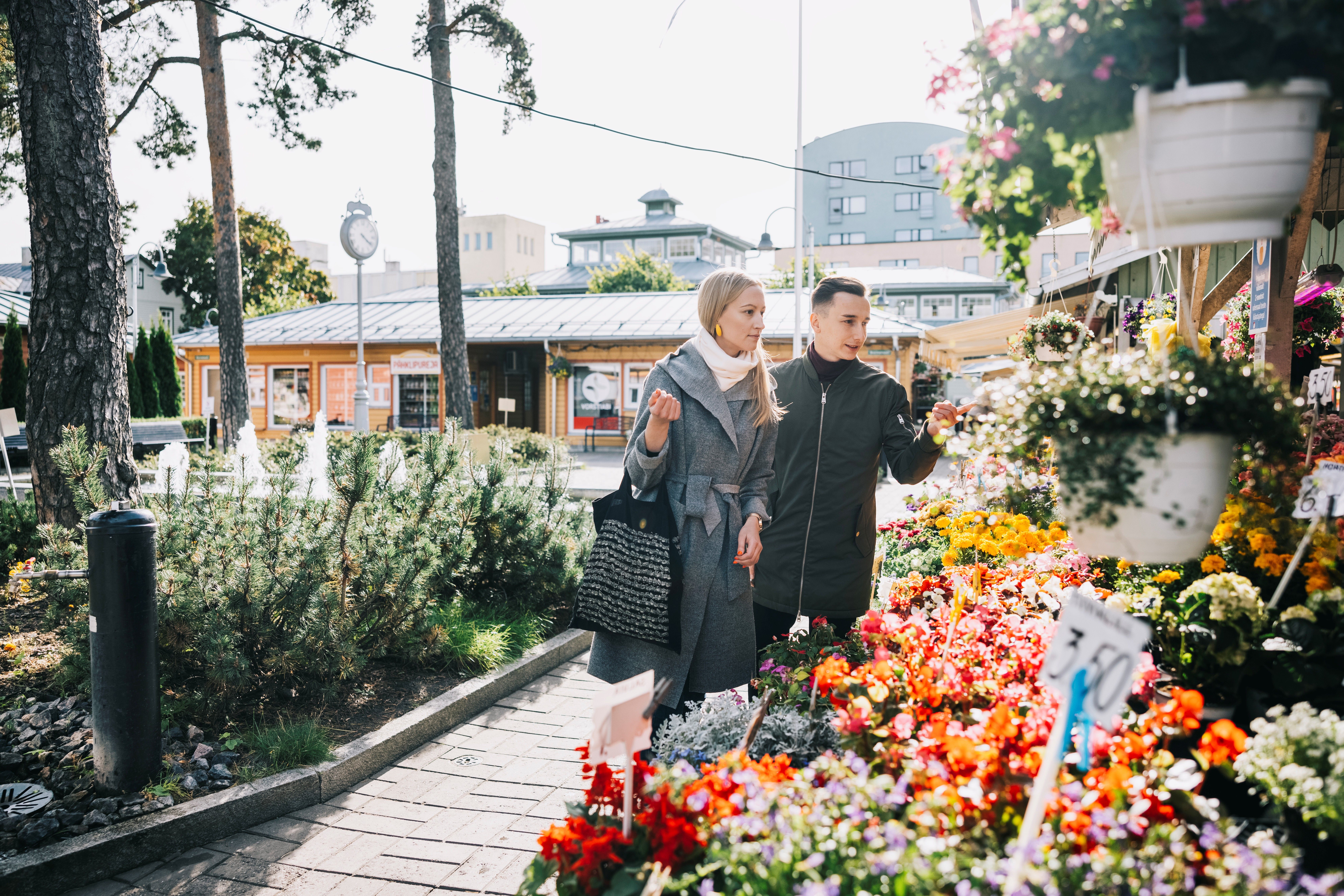 A couple admiring colorful flowers at an outdoor market stall in Nõmme, surrounded by greenery and small shops.