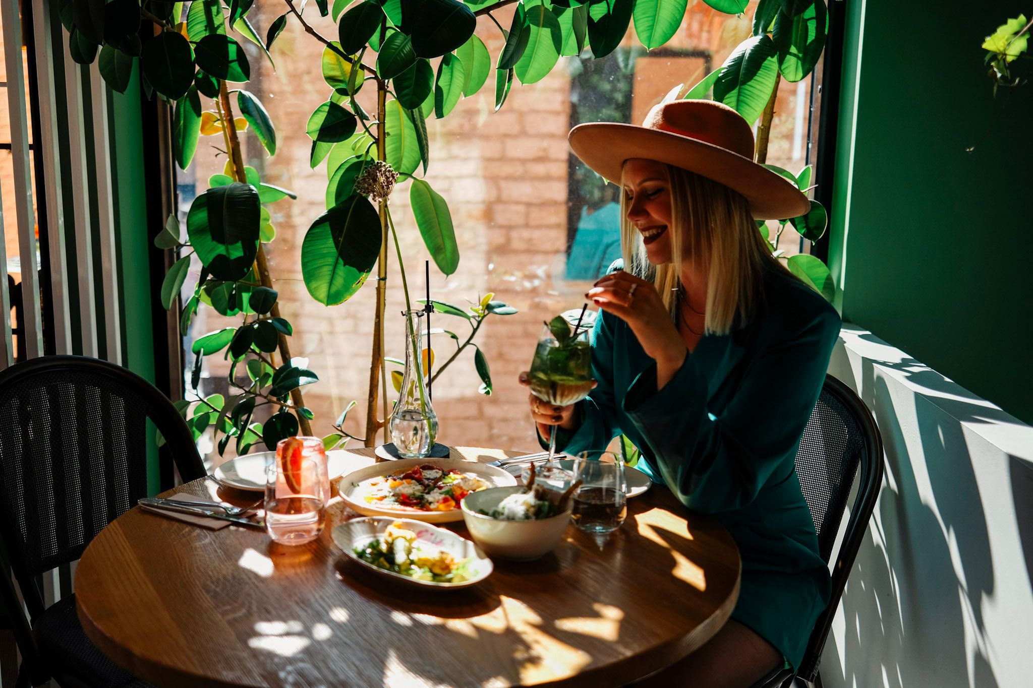 Woman enjoys lunch and a cocktail in a plant-filled caf&eacute; in Tallinn, with sunlight streaming through the window.