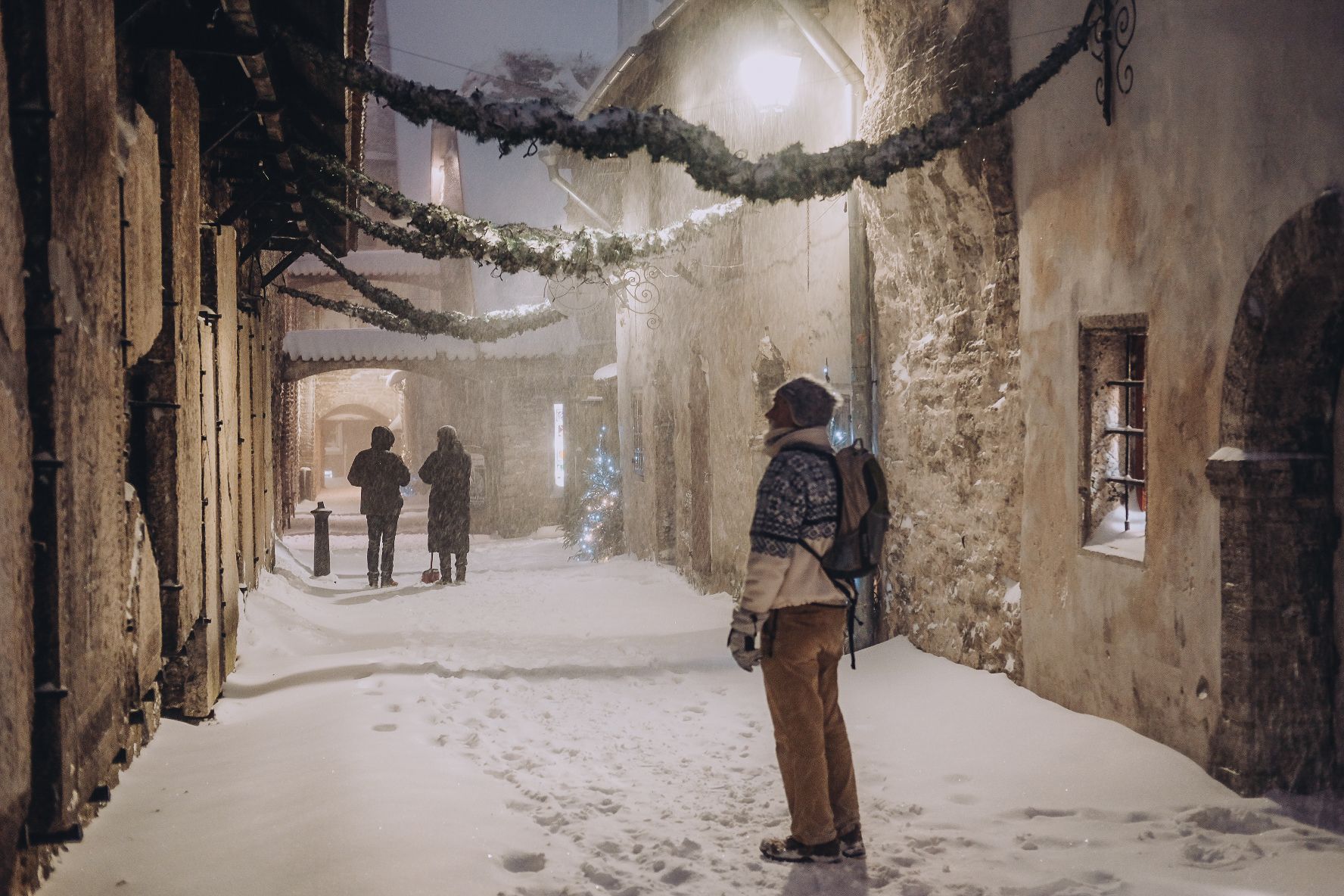 A man wearing a woollen hat and jumper is standing at St. Catherine's Passage in Tallinn, Estonia in the winter. The ground is covered in thick snow.